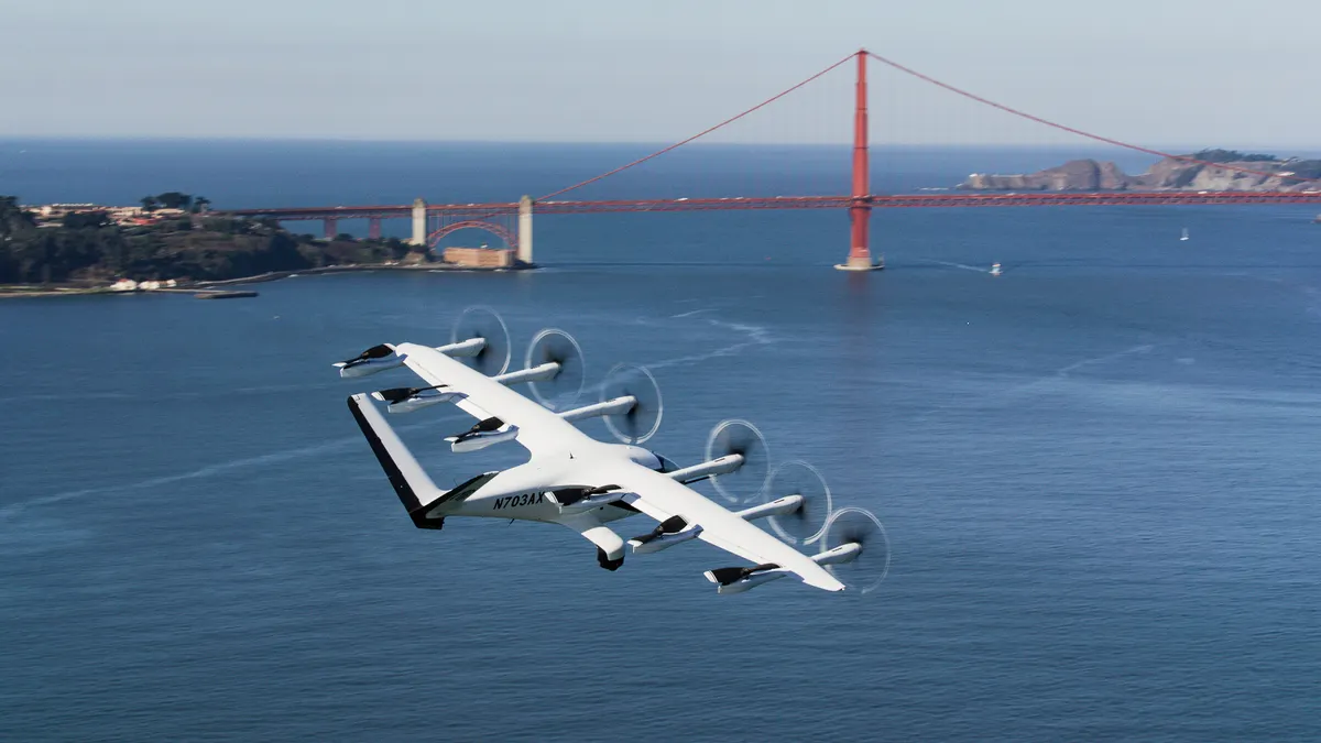 A white airplane with 6 propellers flies over water with a red bridge in the background.