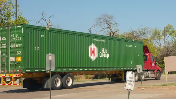 A red semi-truck pulling a large 53-foot green Hub Group shipping container drives past a sign for "Driver Orientation Parking" at the company's terminal.