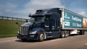 An 18-wheeler drives down a highway. The cab of the truck is dark blue and the trailer has writing on the side that says "McLane. Food & Service drive us."
