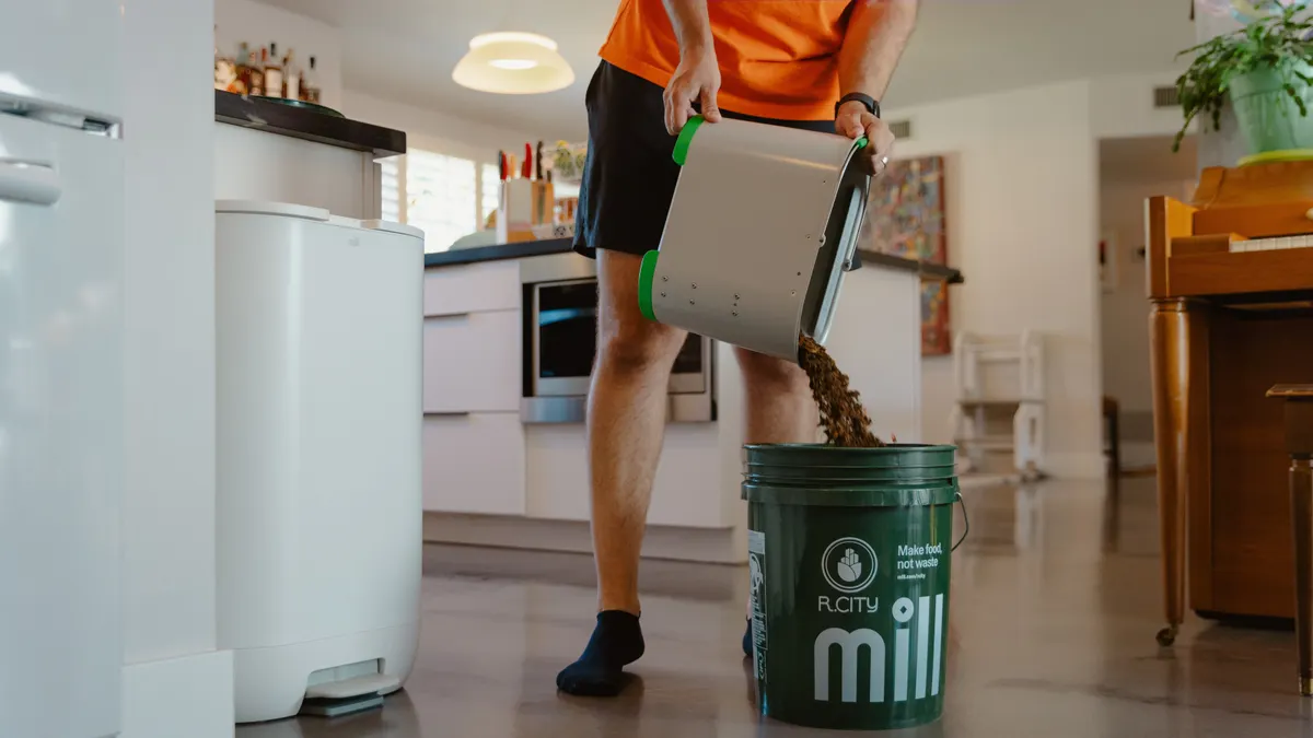 A person standing in a kitchen pours dehydrated food grounds into a 5-gallon bucket labeled with "mill" and "r.city"