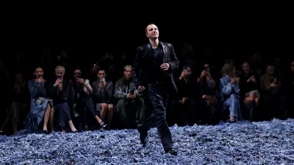 A man walks on a darkened runway that is covered in small pieces of denim as a crowd watches.