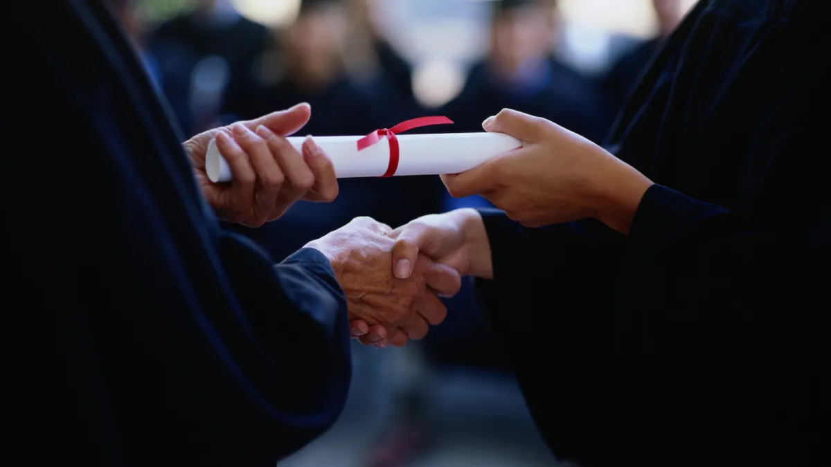 A close up of hands shaking, with one person giving the other a rolled up diploma