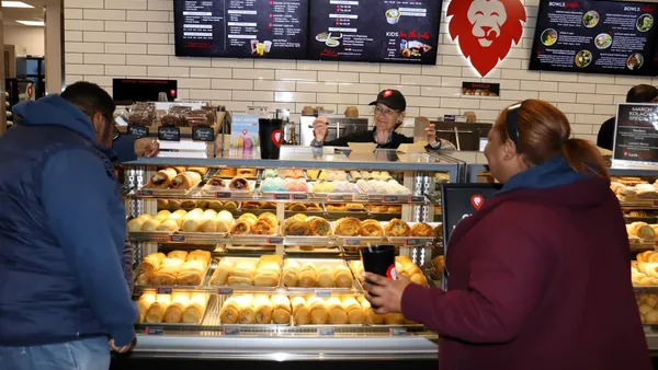 Twp people stand in front of a lit case full of prepared foods, while a third person stands behind the case in an apron. There is a menu board on the wall, too small to read, and a sign showing a red lion licking its lips.
