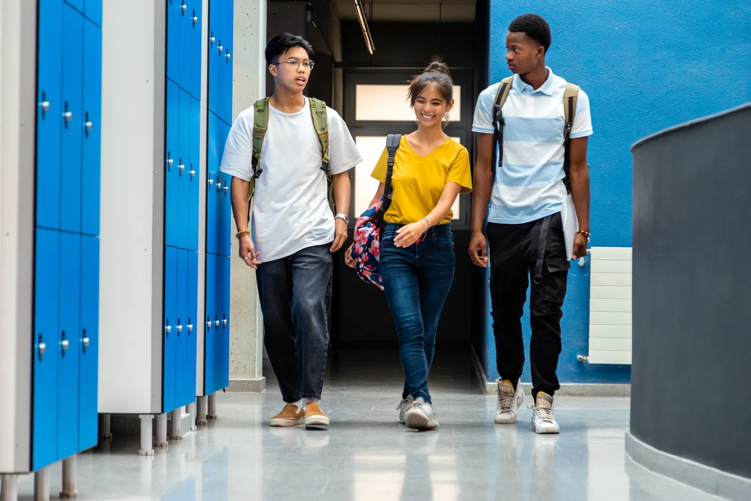 Three high school students walk down a school hallway with locker on the left side wall.