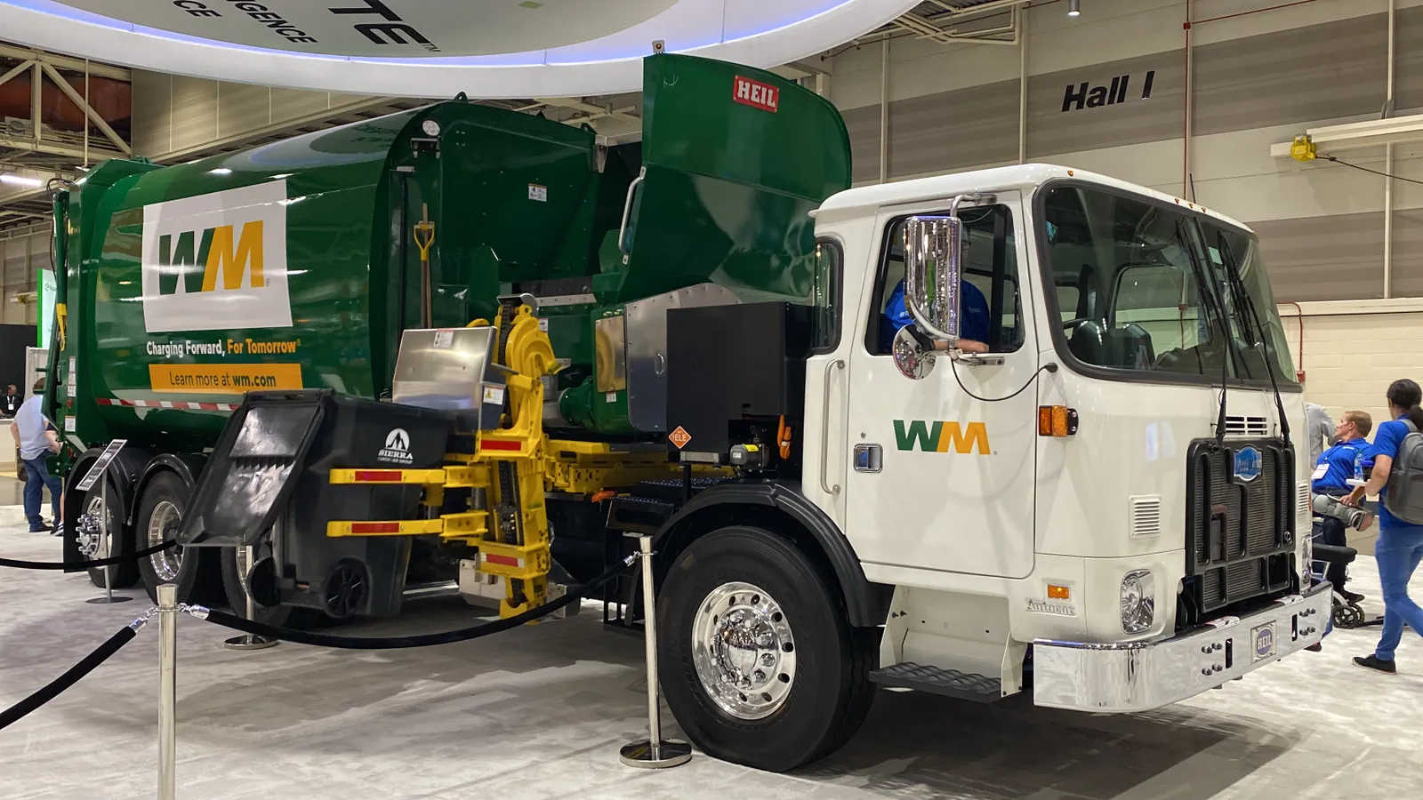 A refuse hauler with WM branding demonstrates side-loading a waste bin