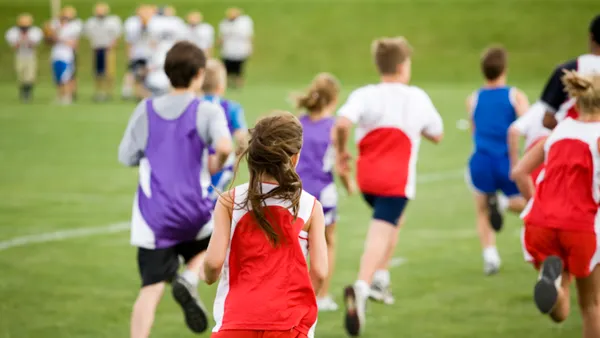 A women runner is in the center of the photo with their back to the camera. Other runners are seen in front of the person and everyone is running on an outside track.