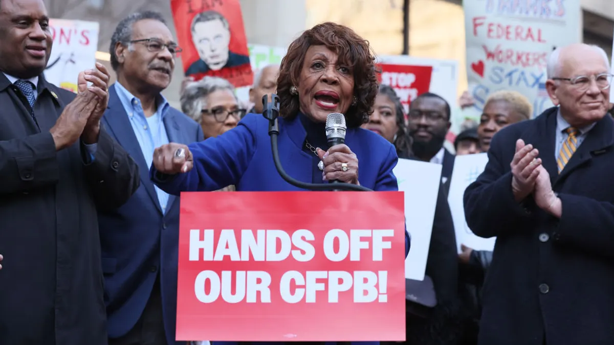 Rep. Maxine Waters speaks into a microphone with a crowd of people behind her, some holding signs. A sign in front of her podium reads "Hands off our CFPB!"