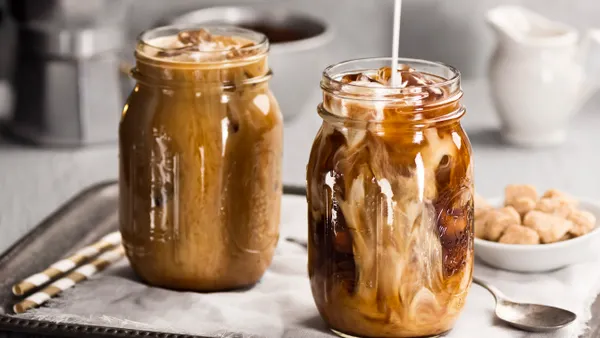 Two iced coffee drinks in glass jars with cream being poured into the one on the right and filtering through. The jars are on a silver tray with a white linen napkin, two straws, a spoon and a bowl.