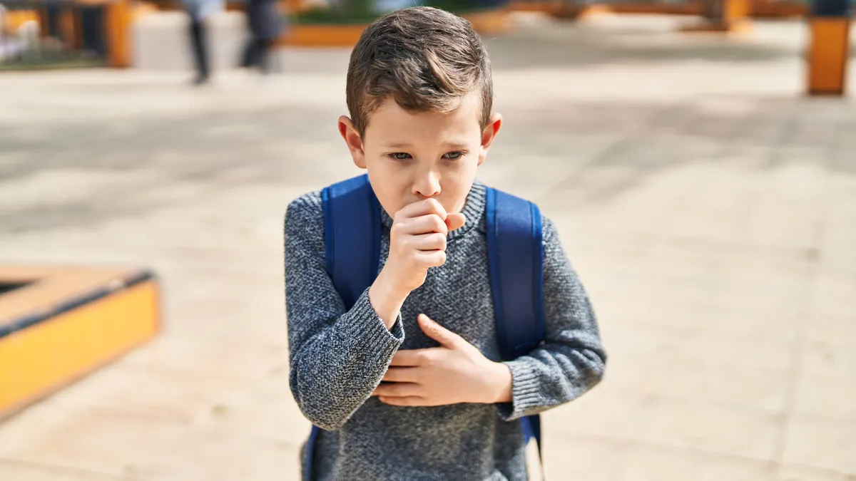 A child in a backpack coughs while walking outside.