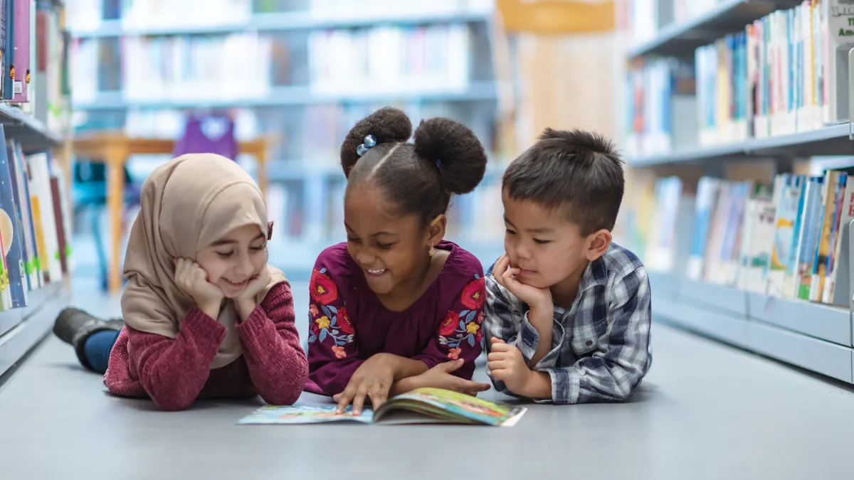 Three young children lie on their stomachs on the floor looking at an open book. There are rows of books on their side of them.