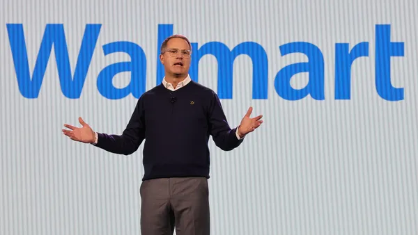 A man speaks while standing in front of a screen displaying "Walmart" in blue letters.