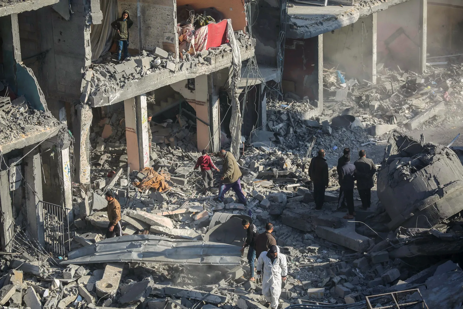 People look and walk around in severe rubble and debris of a half a building facade.