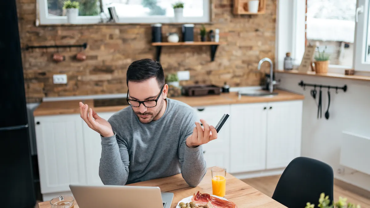 A man is on his laptop and is having problems buying an item online with a credit card. He sits at the kitchen table with breakfast.