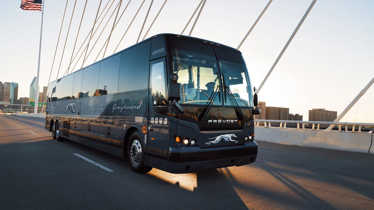 A Greyhound bus on a road with tall buildings in the background.