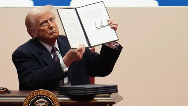 President Donald Trump , seated wearing a dark suit, holds up typed order with his signature on it.