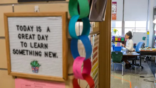 A person is seen sitting at a table in a classroom. The photo is taken from the door of the classroom and near the door is a sign reading "Today is a Great Day To Learn Something New"