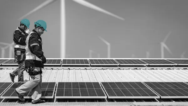 Two workers on a rooftop covered with solar panels, with wind turbines in the background.
