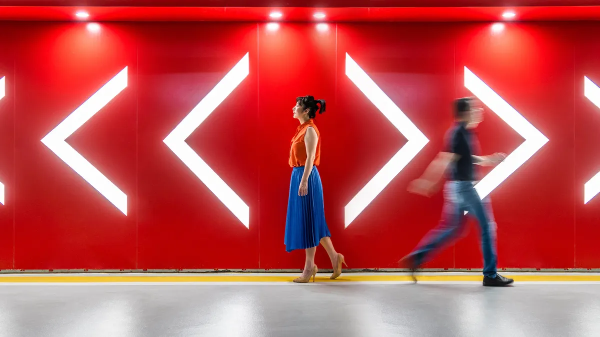 A man and woman walking towards the opposite directions of arrows in front of a red wall.