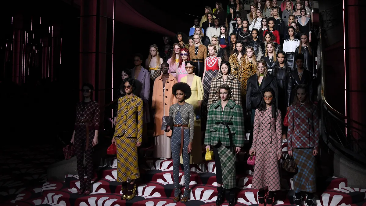 Models stand on a dark runway holding handbags and wearing colorful outfits.