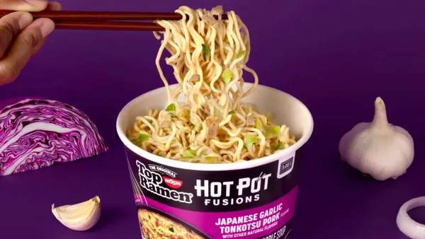A person uses chopsticks to lift noodles out of a packaged cup that reads "Top Ramen Hot Pot Fusions Japanese Garlic Tonkotsu Pork" as chopped cabbage and onions and cloves of garlic lay nearby.