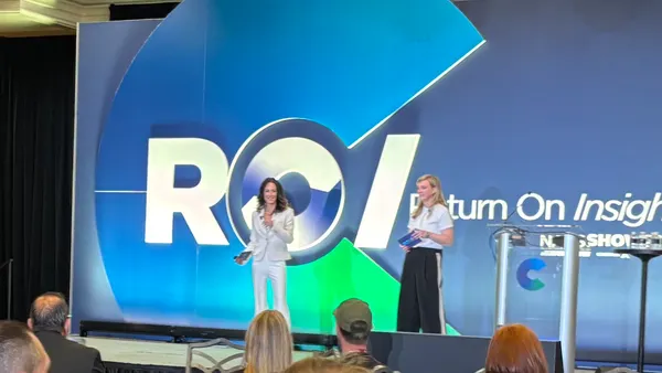 Two people stand on a stage talking to a group of people. Text displayed behind them says "ROI REturn OPn Insight."