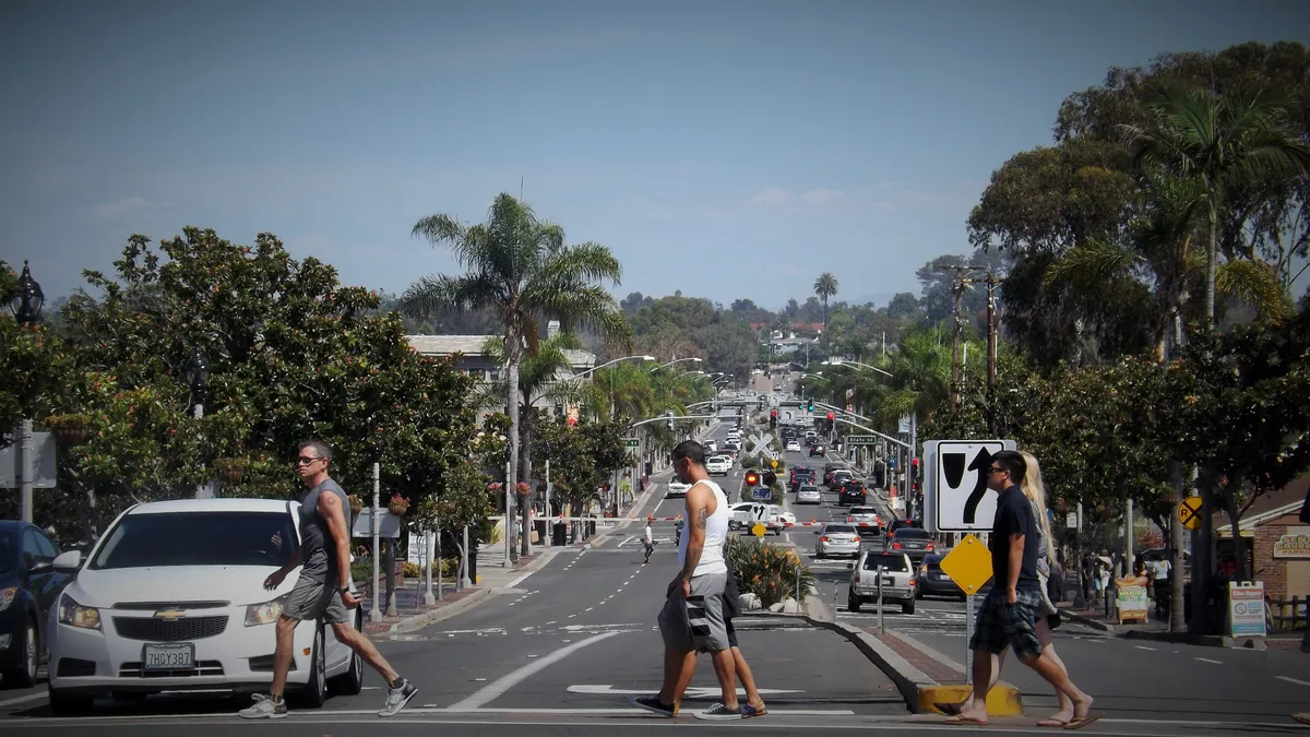 Pedestrians walk across a multi-lane street lined with trees.