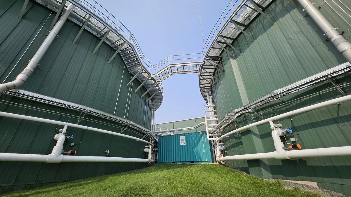 Two massive cylindrical tank buildings are connected by pipes and encircled by catwalks. A building connecting the two has a sign with the "BTS Bioenergy" logo.
