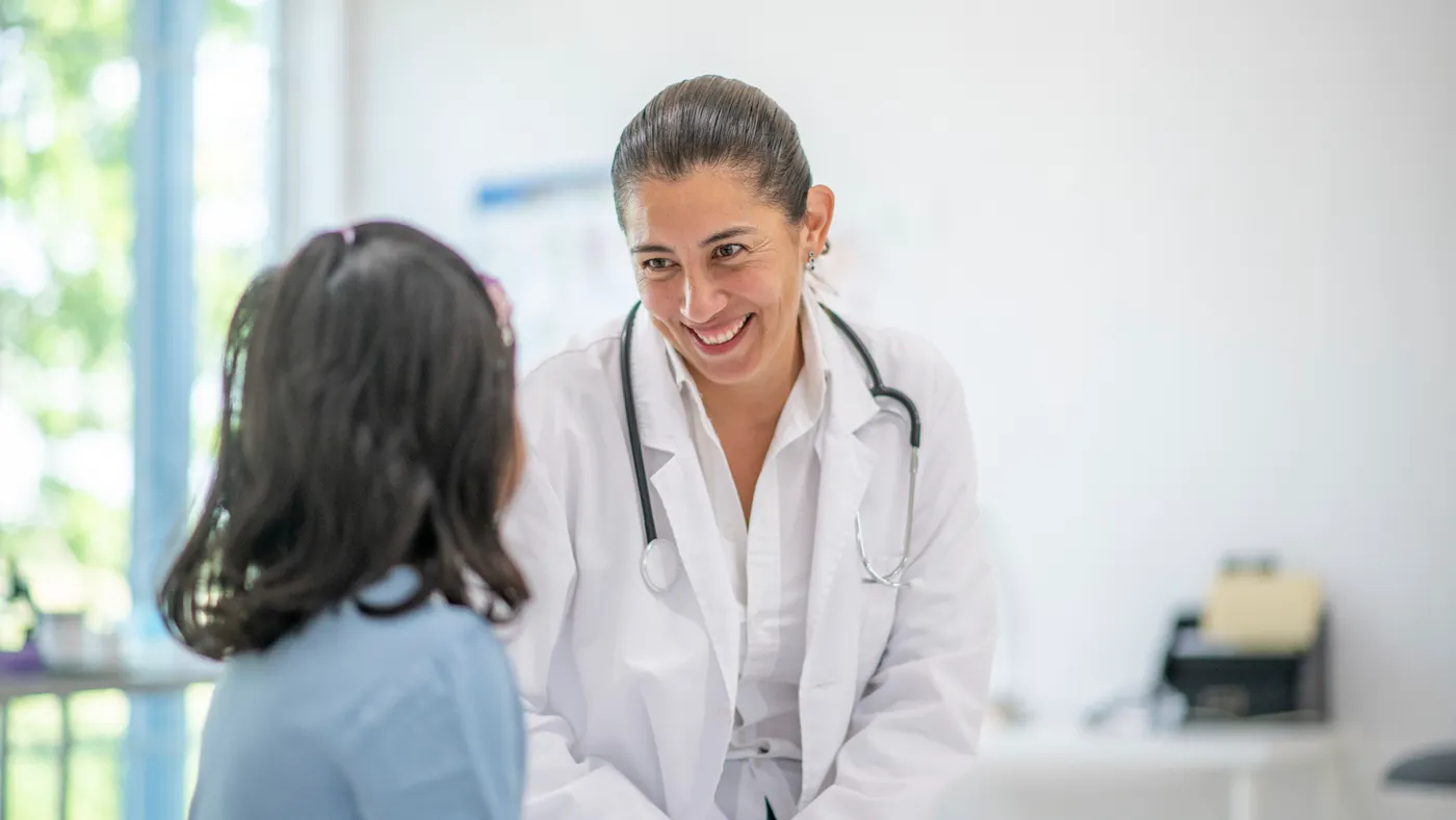An adult in a white lab coat smiles, facing a younger student as they sit on a bench in a room with a window.