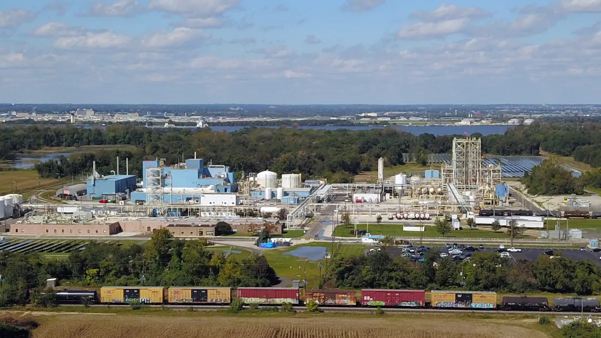 A white manufacturing plant surrounded by green landscape, road and water on a sunny day with white clouds.