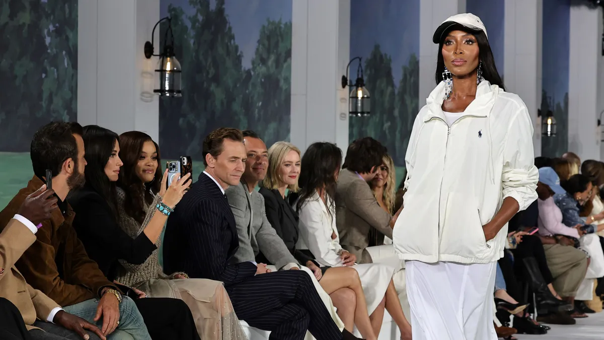 A model, Naomi Campbell, walks down a runway wearing a white jacket as viewers watch from the side.