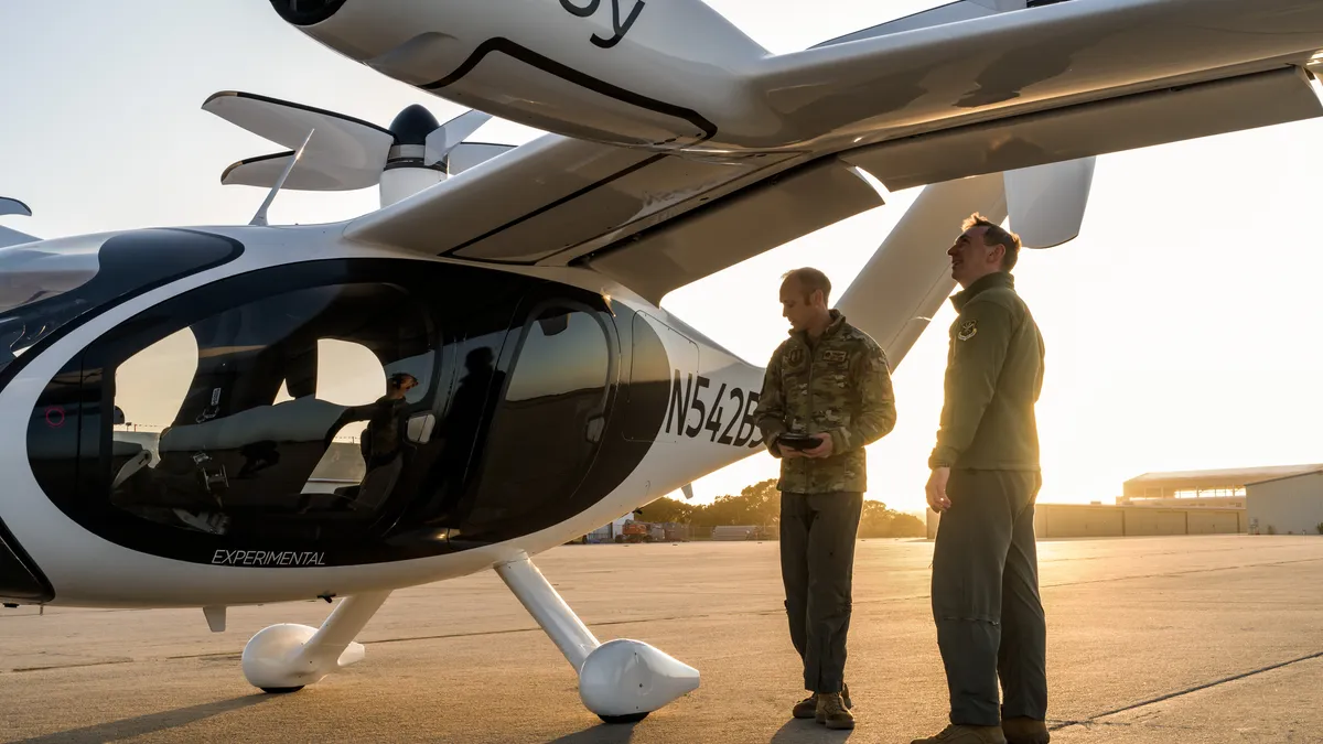 Two men stand aside a futuristic vertican takeoff and landing aircraft on a large concrete pad.