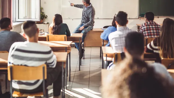 An adult man sits on the edge of a desk in front of a high school classroom as he teaches math from a whiteboard.