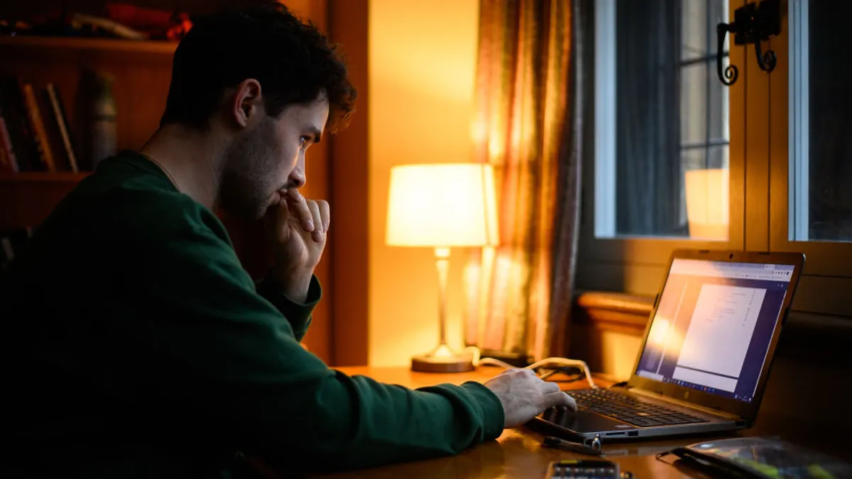 A man in a green shirt sits in a dorm room looking at a laptop screen in lamp light.