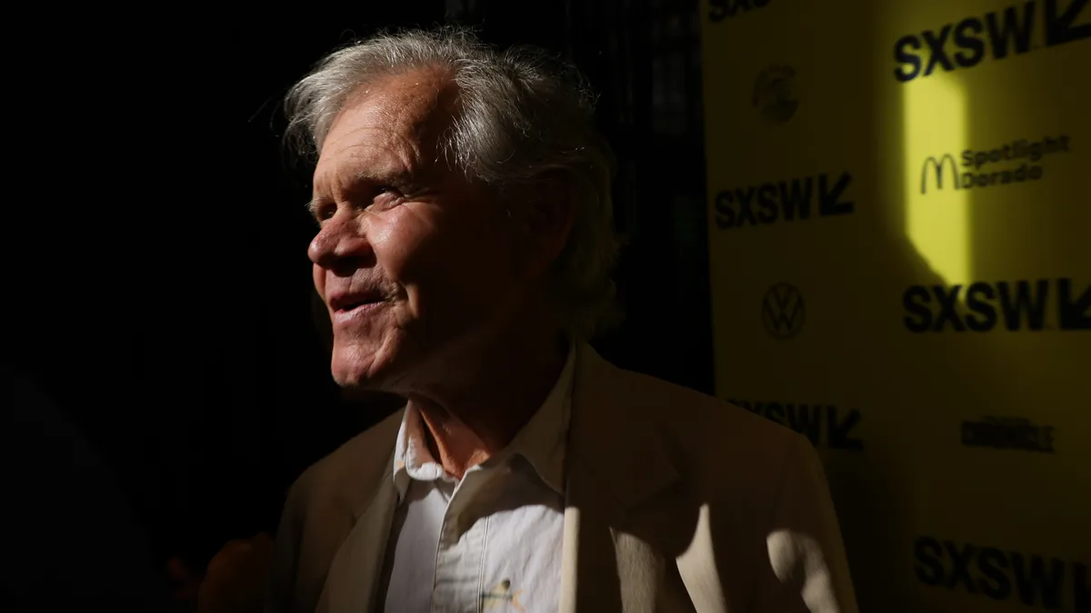 A person with white hair looks to the side against a yellow background.