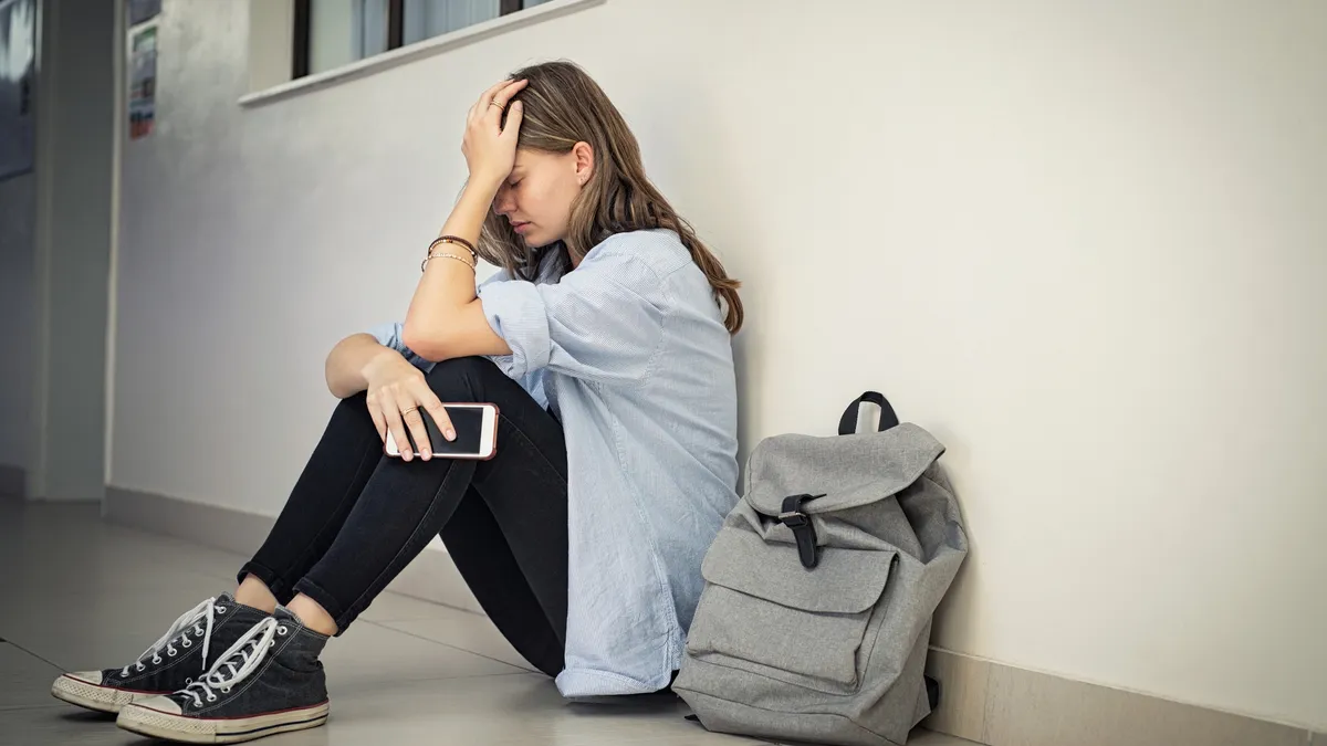 A high school student sits in a school hallway with head in hand and cell phone in other hand.