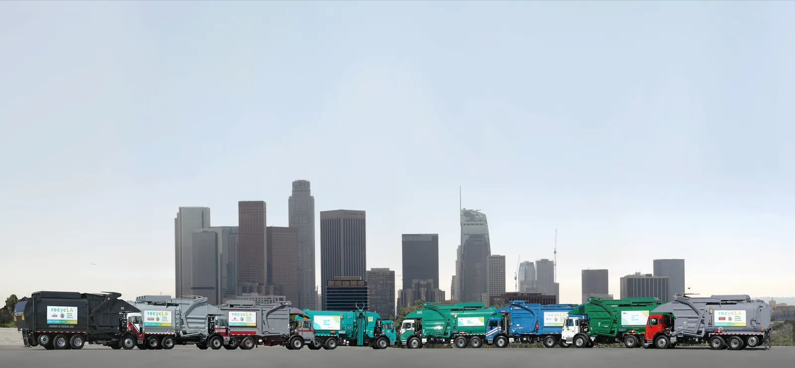 A row of garbage trucks parked in front of the Los Angeles skyline