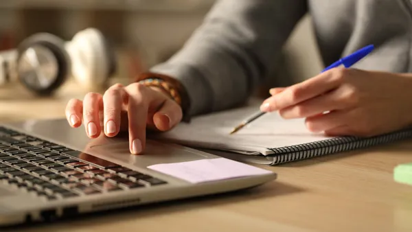 A student writes on a notebook with one hand and scrolls on a laptop with another.