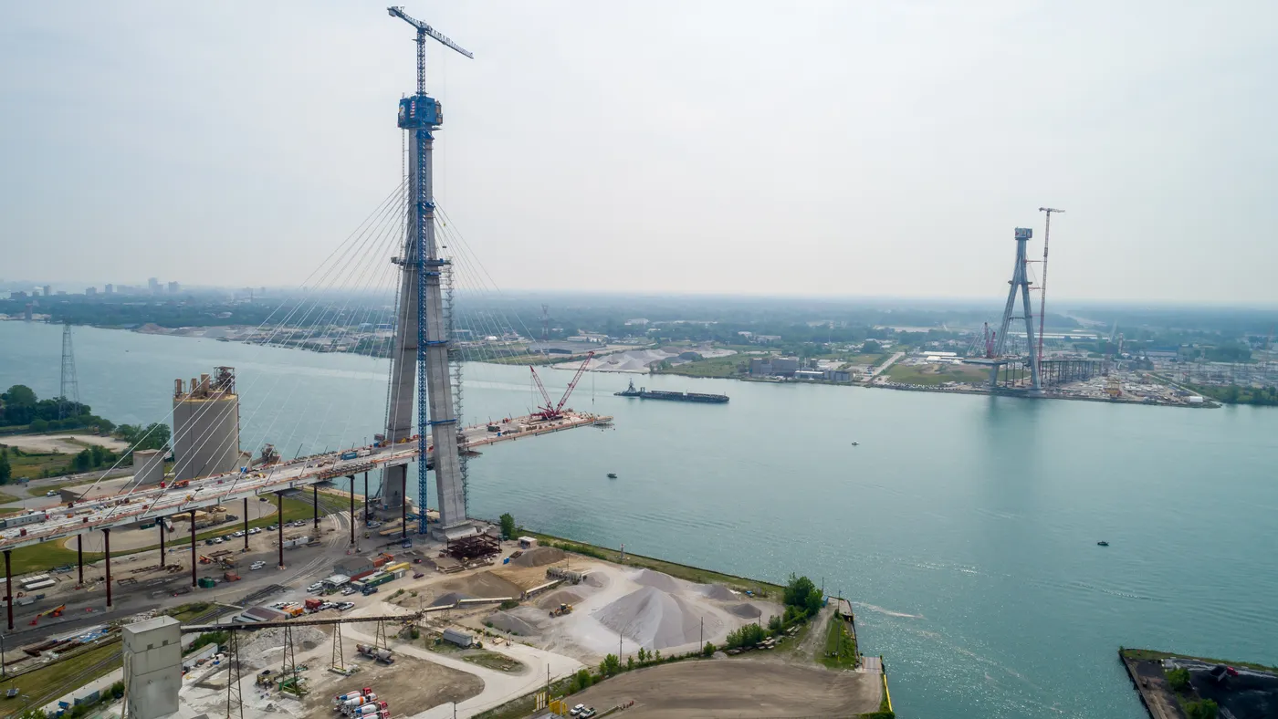 Two bridge towers on either side of a river stand tall, with scaffolding and cranes around them.