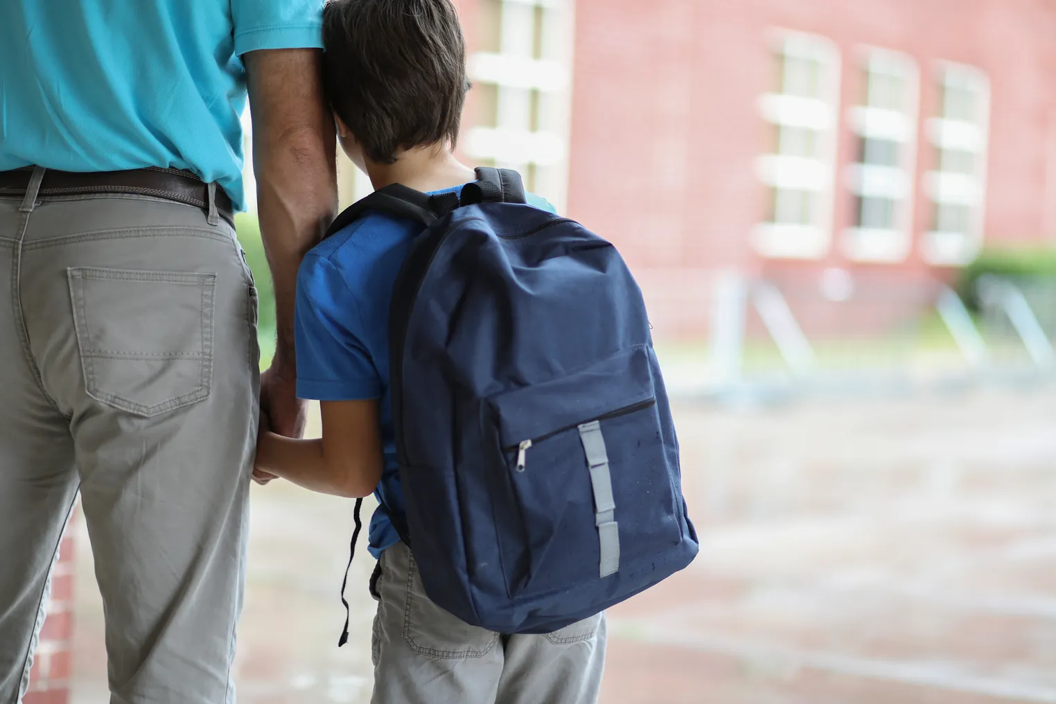 The back of an adult is on the left. The adult is holding the hand of a child carrying a backpack, whose back is also to the camera. The two are standing in front of a building