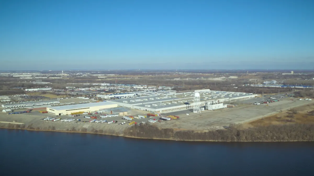 A manufacturing plant sits alongside a river bank. The parking lot shows trailers outside the facility.