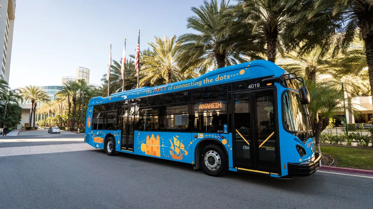 A blue bus parked on a road lined with palm trees, with a modern glass-facade building in the background.