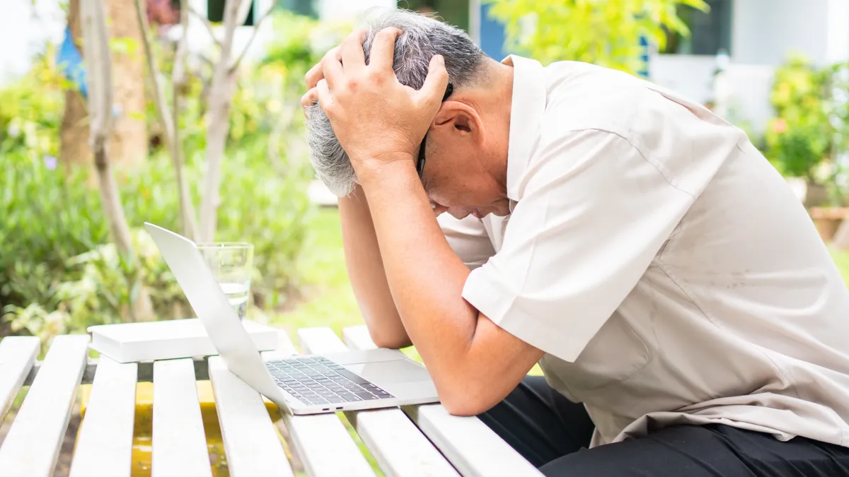 An older man sitting at a laptop holds his head in his hands.