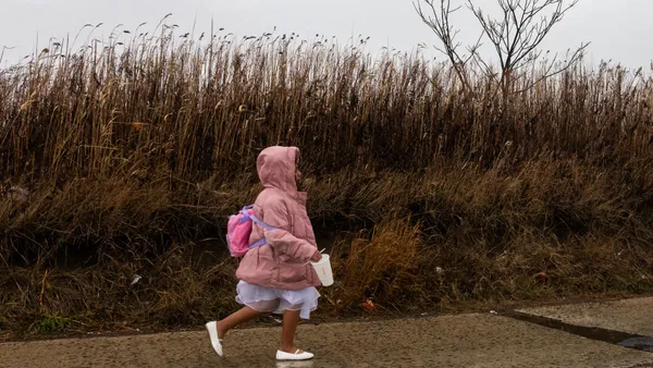 A young student walks outside on a walkway, wearing a pink coat and pink backpack. Tall seagrass is along the walkway.