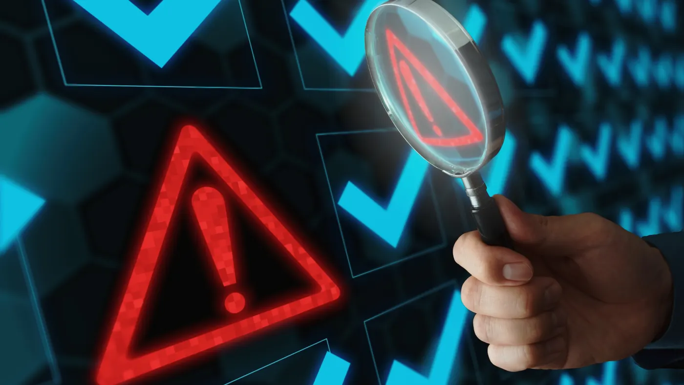 A hand holds a magnifying glass up to a red exclamation point enclosed by a red triangle