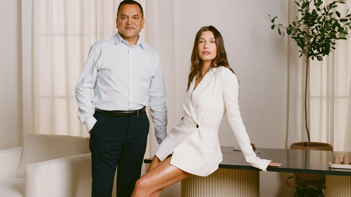 Two people stand together in a cream-colored room by a table.