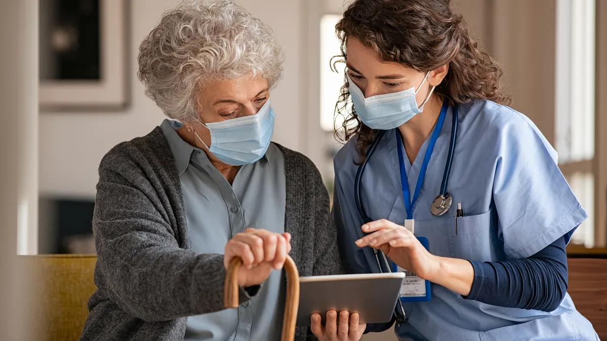 Doctor and senior woman going through medical record on digital tablet during home visit wearing face mask.