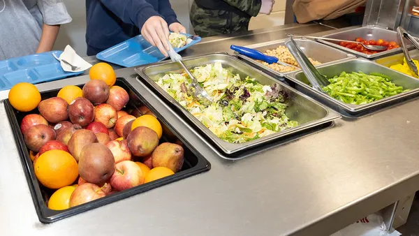A photo of a school lunch buffet of salad, apples and vegetables. Students' hands and arms can be seen filling their plates with food.