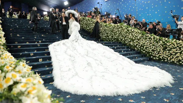 A person in a large white hat and long, flowing white flowing cape stands atop a blue staircase.