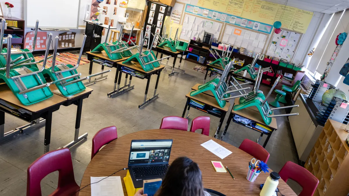 A teacher sits at a desk while virtually instructing students through a laptop. The teacher is in an empty classroom with rows of chairs flipped upside down on top of students' desks.