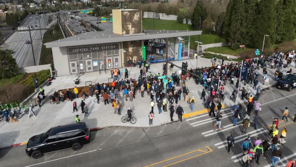 An aerial view of a train statin alongside a highway with crowds of people walking toward the gray station building.